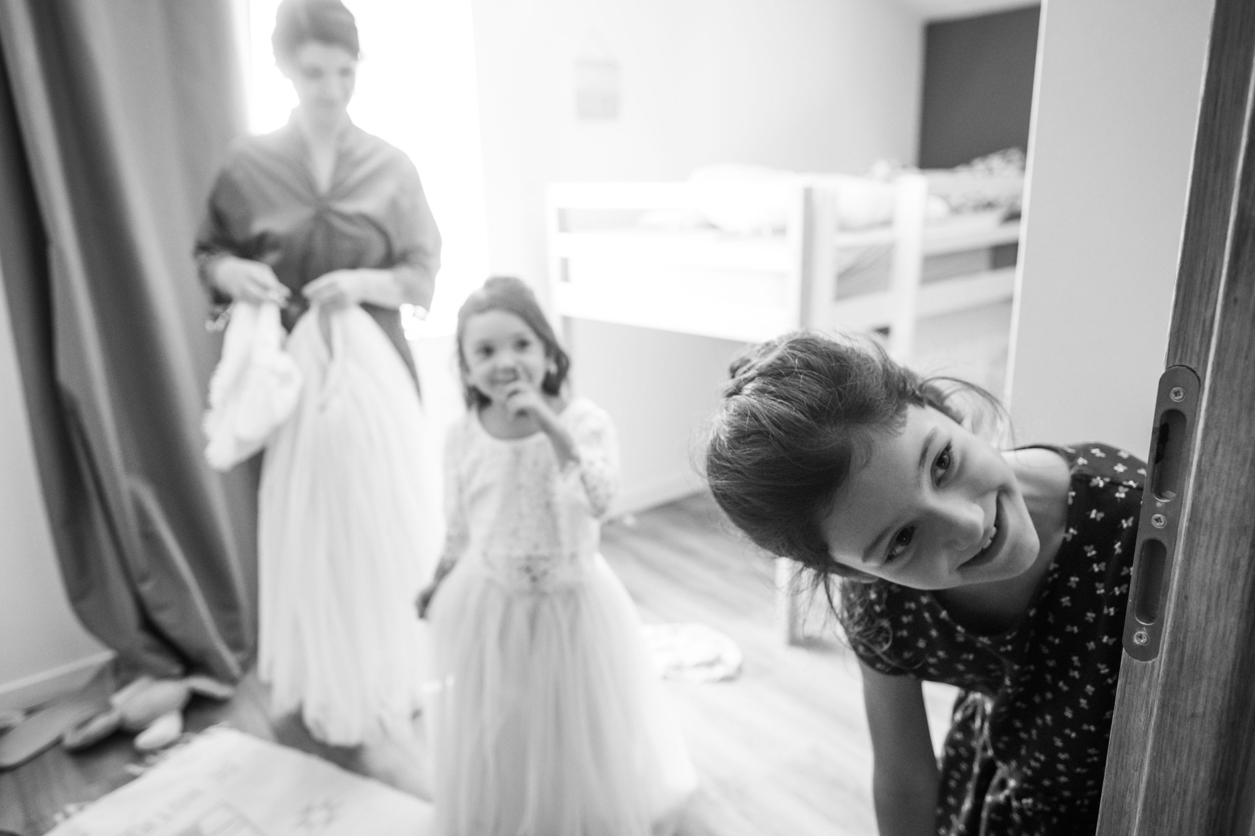 Une amie de la mariée passe la tête par la porte en souriant, une petite fille en robe de cortège derrière elle