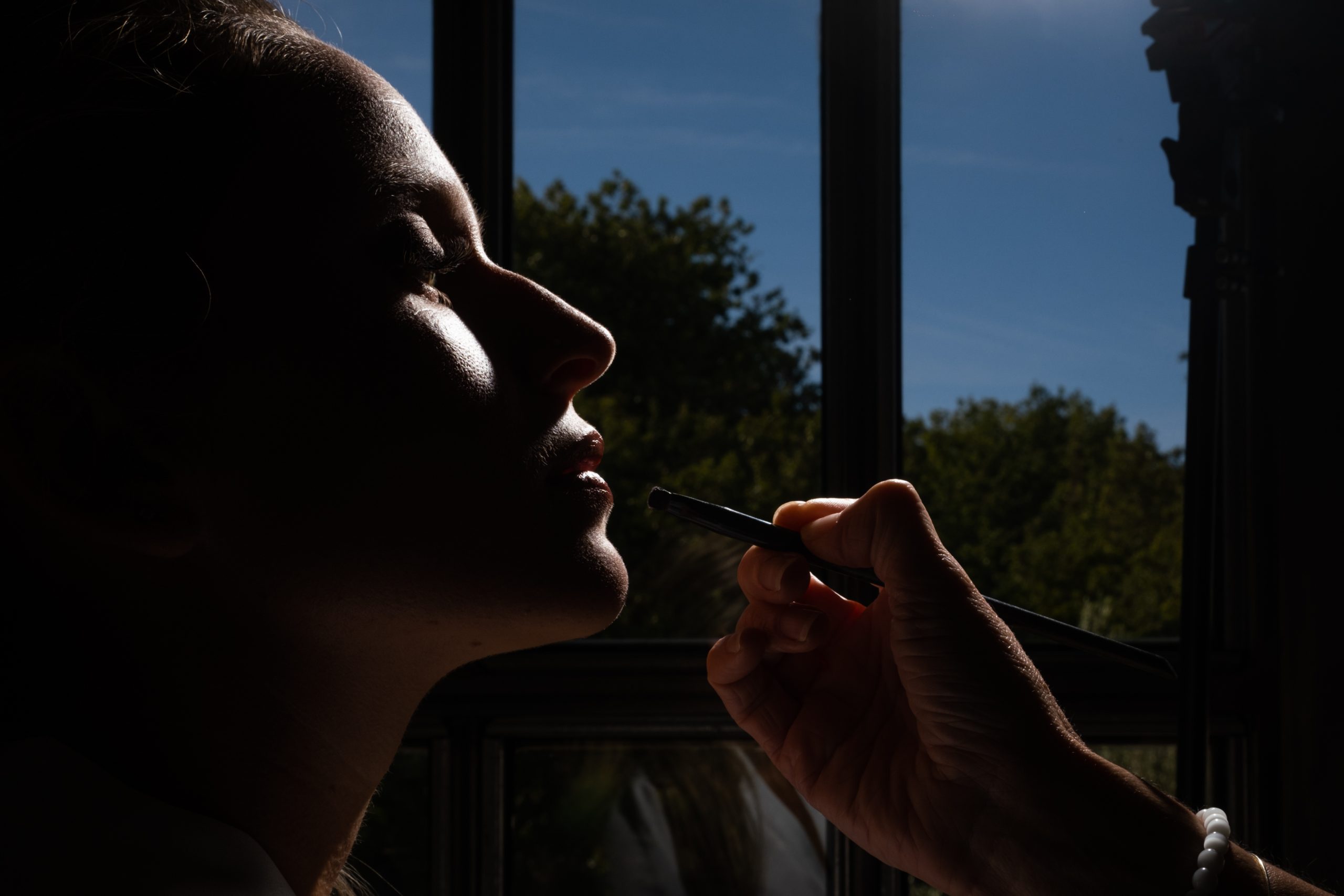 Maquillage des lèvres de la mariée sculpté par un coup de flash, en contre-jour devant la fenêtre, préparatifs de mariage en Drôme