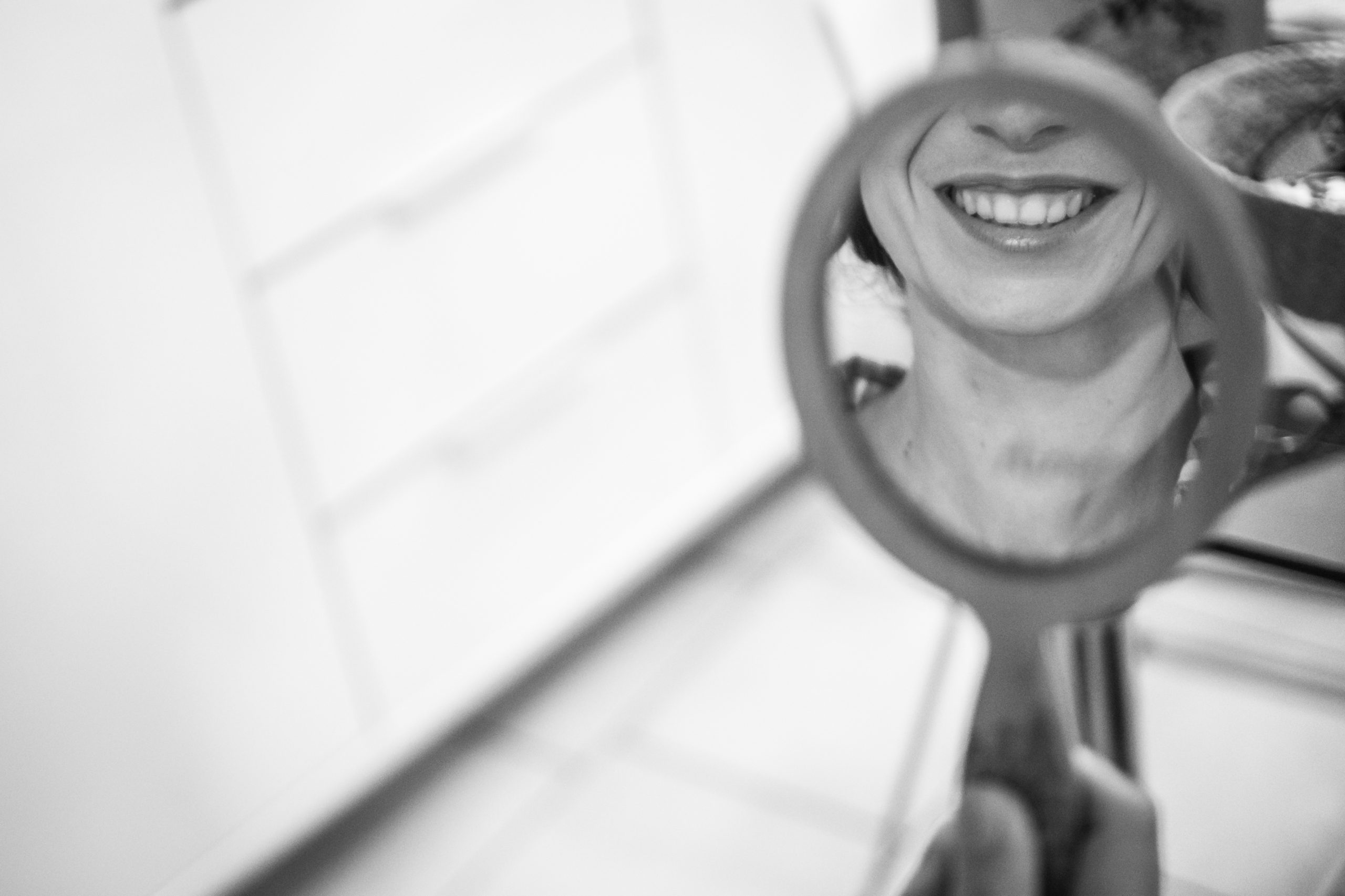 Sourire de la mariée dans le reflet d'un petit miroir à main pendant les préparatifs, en noir et blanc
