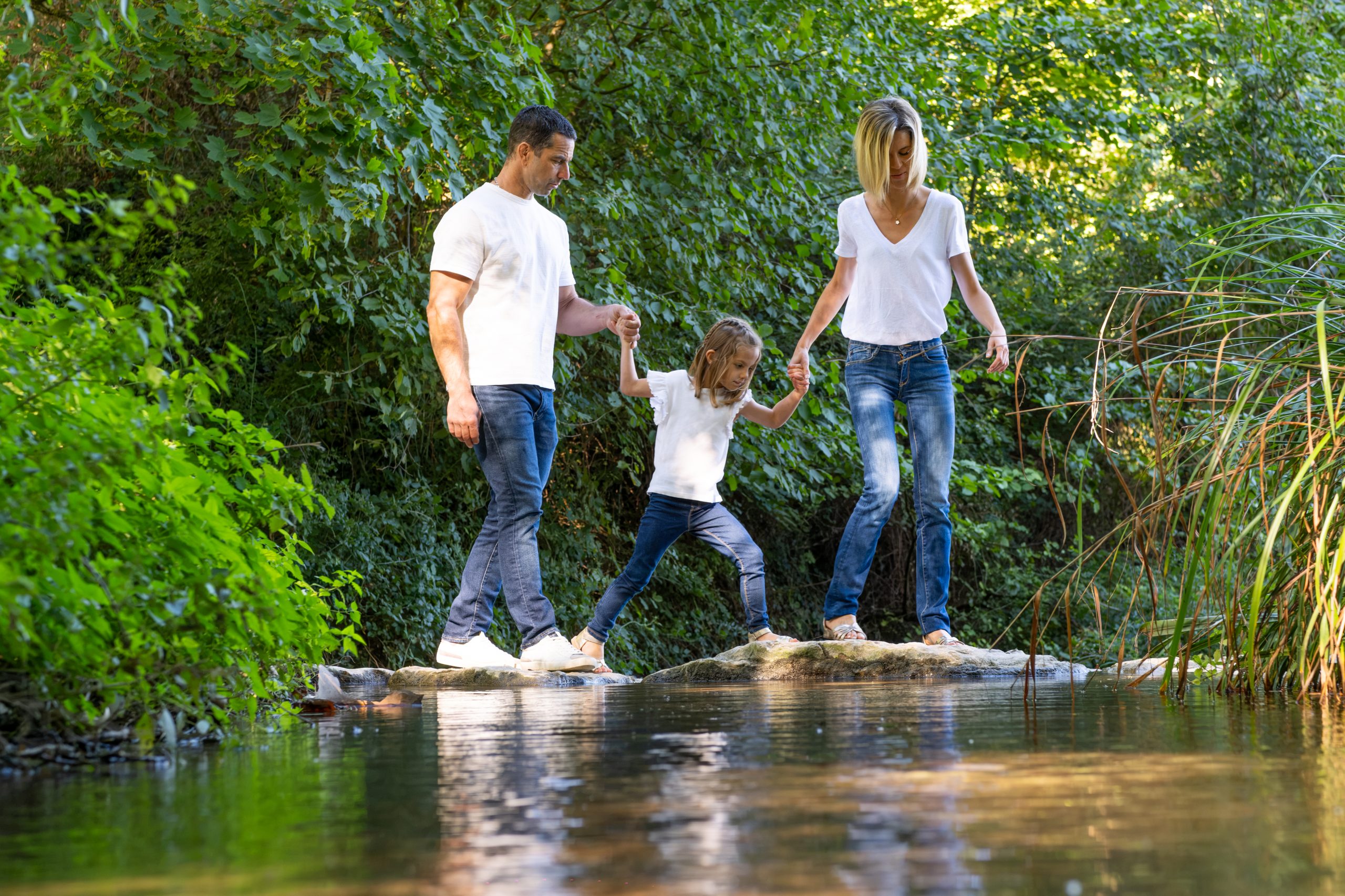 parents et leur fille traversant une rivière main dans la main, séance photo en famille en Drôme